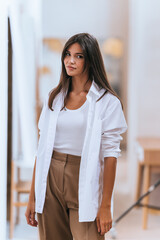 Confident young woman in white shirt over tank top, with brown trousers, indoor