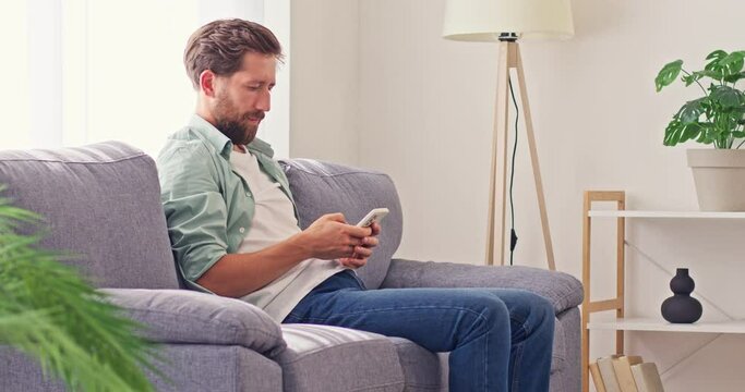 Happy man, sitting on comfortable sofa at home, uses modern mobile phone, scrolls news feed, then puts device away, puts hands behind head, enjoys free time, feels relaxed and satisfied