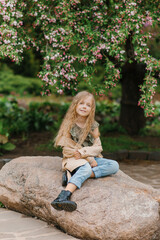 Caucasian cute baby girl with long wavy blonde hair in a trench coat sitting on a stone in spring in a blooming park