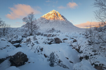 A frozen mountain river and waterfall at the foot of a snow-capped mountain with a morning sunlit peak. Etive Mor Waterfall. Scottish Highlands, Scotland
