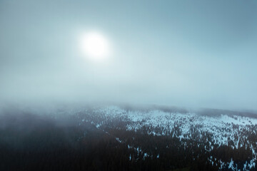 Aerial view of snowy mountain range in cloud with sun shining through during frosty day. Moody...