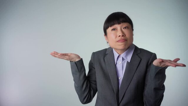 Portrait of a confused Asian business woman shrugging shoulders isolated over grey background. Woman with short haircut, spreads hands sideways, smirks face, feels doubt while makes choice