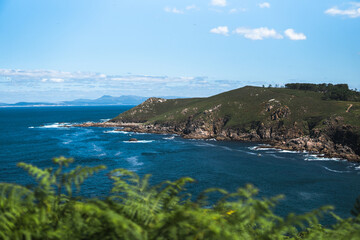 Naklejka premium Captivating Ons Island scene: lush green ferns in the foreground, azure sea, rocky cliffs, Pontevedra estuary, and a summer sky with scattered clouds from the Buraco do Inferno path.