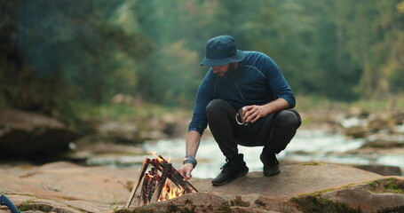 A traveler drinks tea from a metal mug near a campfire during a tourist hike in the mountains