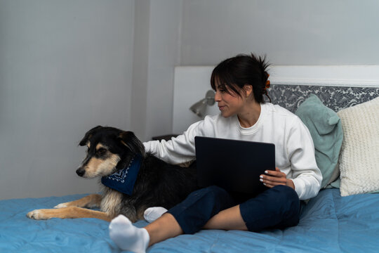 A woman comfortably works from home on her bed with a laptop, accompanied by her loyal dog wearing a blue bandana