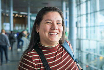 Young Latina woman at the airport, excited for the trip, with a wide and radiant smile, eager for the holidays, living with a positive attitude, with two bags hanging from her shoulders. Close-up shot