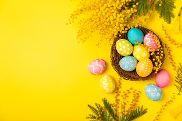 Easter eggs in the nest with mimosa flowers on a bright yellow background. Easter celebration concept. Colorful easter handmade decorated Easter eggs. Place for text. Copy space.