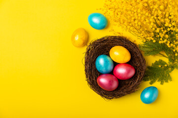 Easter eggs in the nest with mimosa flowers on a bright yellow background. Easter celebration concept. Colorful easter handmade decorated Easter eggs. Place for text. Copy space.