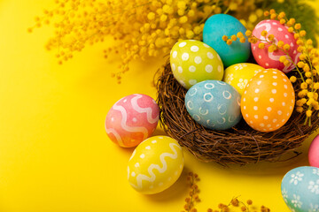 Easter eggs in the nest with mimosa flowers on a bright yellow background. Easter celebration concept. Colorful easter handmade decorated Easter eggs. Place for text. Copy space.