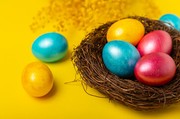 Easter eggs in the nest with mimosa flowers on a bright yellow background. Easter celebration concept. Colorful easter handmade decorated Easter eggs. Place for text. Copy space.