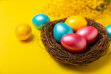 Easter eggs in the nest with mimosa flowers on a bright yellow background. Easter celebration concept. Colorful easter handmade decorated Easter eggs. Place for text. Copy space.