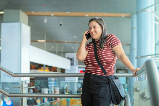 Young woman talking on her cellphone at the airport, with a shoulder bag, stairs, big smile, and excitement for her upcoming airplane trip