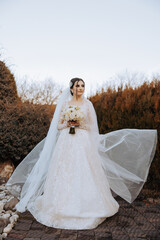 A brunette bride in a voluminous white dress holds a bouquet, poses. The veil is thrown into the air. Beautiful hair and makeup. Wedding photo session in nature. Long train of the dress