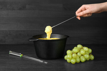 Woman dipping grape into fondue pot with melted cheese at black wooden table, closeup