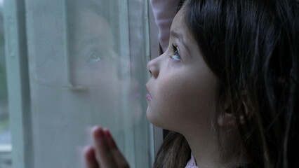 Sad little girl leaning on glass window with pensive expression, melancholic emotion of child struggling with loneliness and solitude