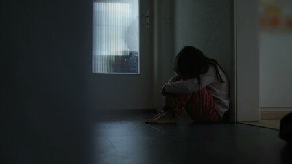 Sad lonely child seated at home corridor in moody gloomy dark scene covering face in anxiety and...