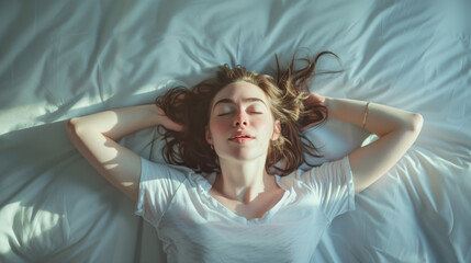 Fototapeta premium Top view of a relaxed young woman lying on bed with hands behind head, basking in morning sunlight.