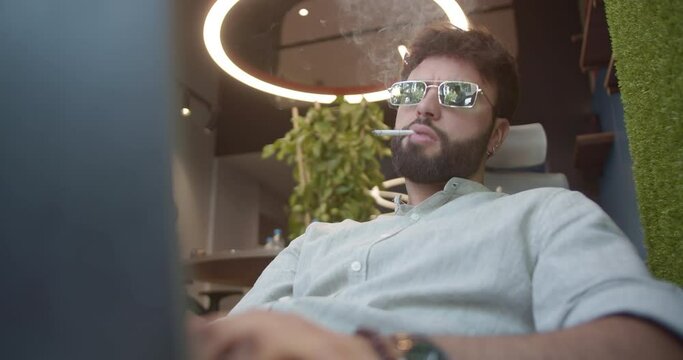 A shot of handsome, male business person smoking cigarette working on his individual tasks using lap top while lying on a sofa at the office.