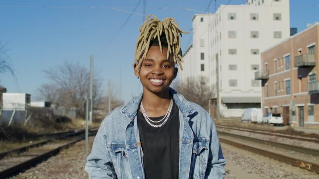 genderfluid person smiles at the camera while standing on train tracks in an urban area