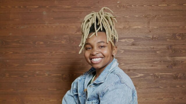 genderfluid person with blonde dreadlocks smiles at the camera with arms crossed in fron of a wooden backdrop wall
