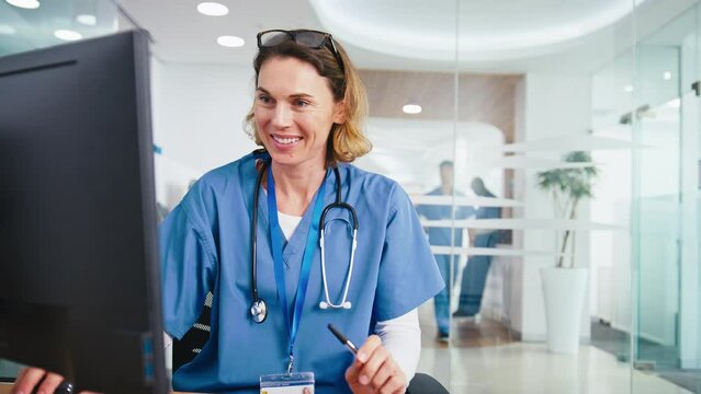 Female Doctor Or Nurse Wearing Scrubs Working At Computer In Hospital Office
