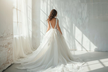 Fototapeta premium Elegant bride from behind showcasing white wedding gown with train against a bright window. Concept of wedding fashion, bridal wear, sophisticated design, and tranquil bridal scene.