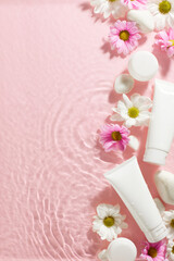 Natural radiance: the essence of spring skincare. Top view vertical shot of skincare tubes, white stones, mix of white and pink flowers on rippled pink water background with space for beauty promo