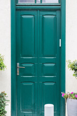 Classic European deep green door, with glass panel and brass handle, flanked by potted pink flowers and greenery.
