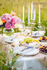 Romantic sunset wedding table decor for a couple with vintage chandelier, burning candles, snacks, white wine, fruits, ranunculi flowers. Summer vibes, field or meadow, outdoors event