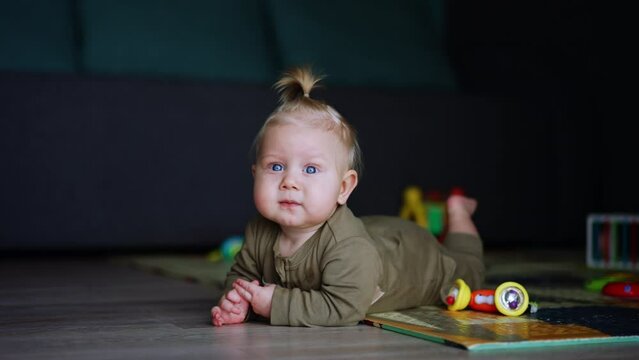 Little baby on bodysuit lies on the floor indoors. Blond child is trying to crawl by the floor and cries.