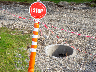 A stop sign in a park in front of an open manhole. Danger lies ahead. A lopsided sign. There is no...