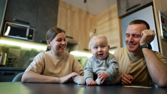 Sweetest baby boy with plump cheeks sits on the table. Loving parents are beside their lovely son. Family in the kitchen. Low angle view.