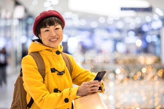 Portrait of smiling Asian woman in yellow coat standing in shopping center and using smart phone.