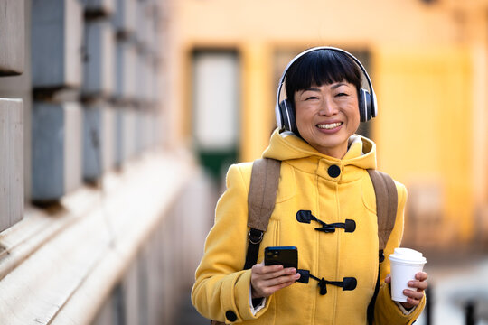 Portrait of smiling Asian woman in trendy coat standing in the street and holding cup of coffee.