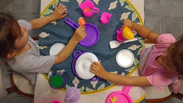 Twin girls playing cooking with colorful toys on table at home, top view