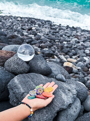Healing crystal and hand holding rainbow gemstones, chakra stones, on grey volcanic ocean shore.