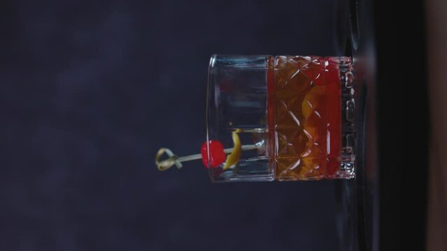 Old Fashioned cocktail drink chilean version with bourbon whiskey, sugar, ice, bitter dashes, maraschino cherry in dark background and selective focus, vertical video