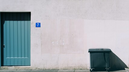 Trash bin in front of a building wall with a blue door illuminated by sunlight.