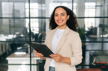 Portrait of confident beautiful curly haired business woman, corporate manager, dressed in a formal wear, standing in a modern creative office, looking at the camera, smiling friendly