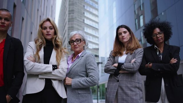Low angle in motion female entrepreneurs in suits from empowered women business in city. Group of five serious coworkers of diverse races and ages looking confident at camera with arms crossed outdoor