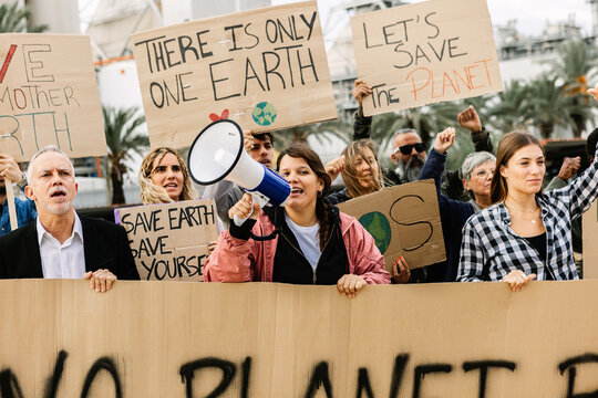 Crowd of diverse protestors demonstrating against climate change in front of factory. Global warming concept with activist holding placards and raising fists. - Powered by Adobe