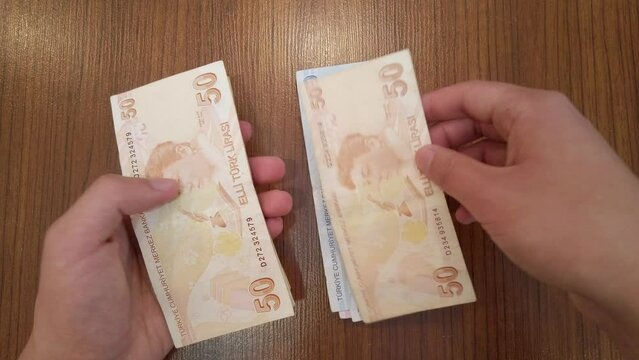 A teenager counting random amount of Turkish lira banknotes with portraits of famous Turkish figures from science, literature and music. He puts banknotes on wooden table, then rearranges them neatly.