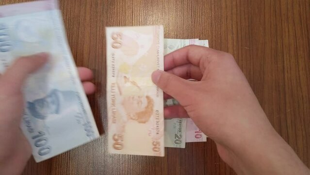 A teenager sorting each banknote of Turkish lira by value, with portraits of famous Turkish figures from science, literature, and music on top. He puts sorted banknotes on wooden table in neat stacks.