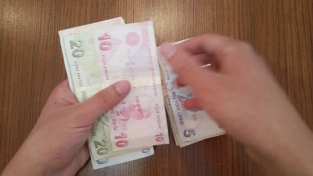 A teenager counting random amount of Turkish lira banknotes with portraits of famous Turkish figures from science, literature and music. He puts banknotes on wooden table, then rearranges them neatly.