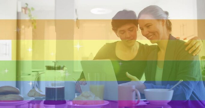 Animation of pride text, rainbow stripes over happy caucasian lesbian couple using tablet at table