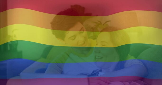 Animation of pride rainbow flag over happy caucasian lesbian couple using laptop in bed