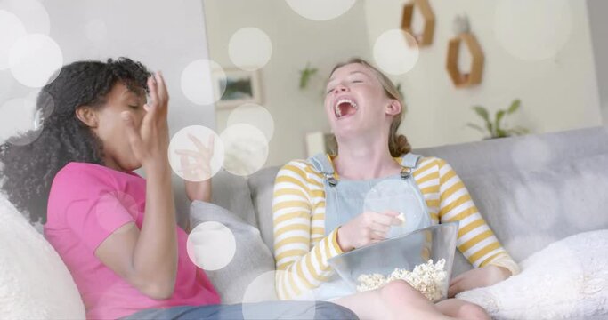 Animation of white light spots over two diverse teenage girls laughing and playing with popcorn