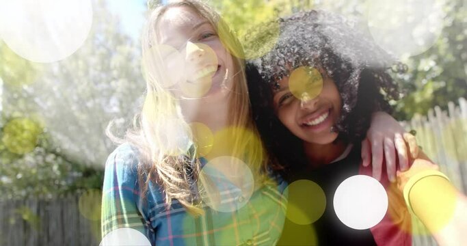 Animation of yellow and white light spots over two happy diverse teenage girls embracing in garden