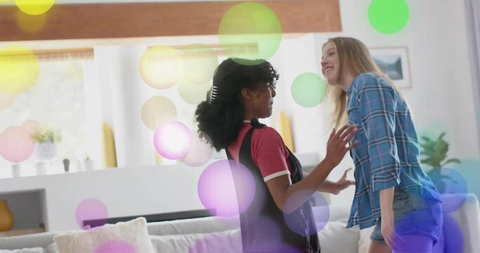 Animation of colourful light spots over two diverse teenage girls dancing at home