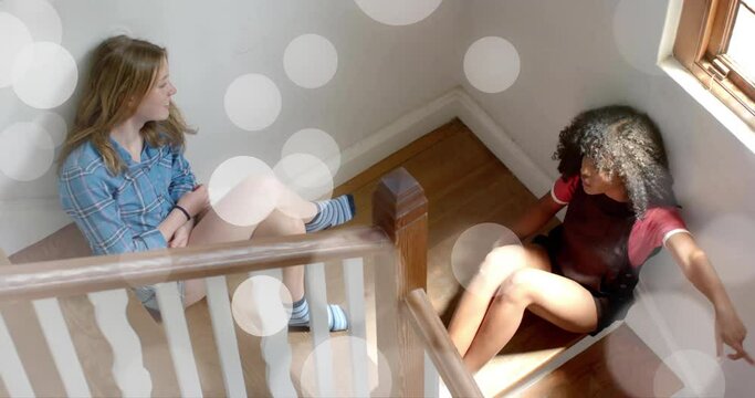 Animation of white light spots over two diverse teenage girls siting on stairs talking at home